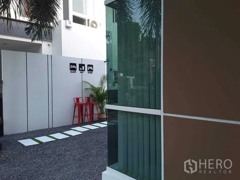 บ้าน ให้เช่าในLam Luk Ka, Pathum Thani - สวนข้างบ้านพื้นโรยกรวด ทางเดิน และเก้าอี้บาร์สีแดงติดผนังกันสายตา