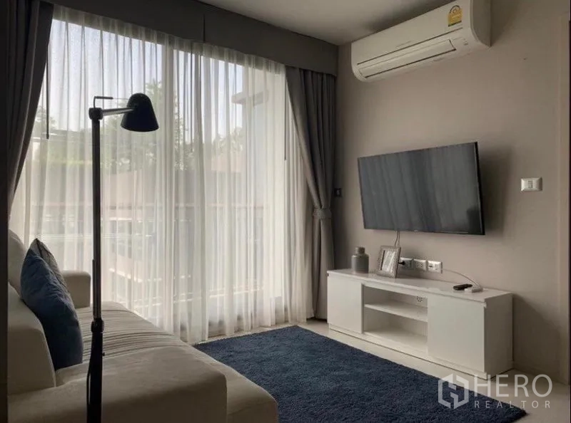 คอนโด ให้เช่าในKhlong Toei, Bangkok - มุมนั่งเล่นมีทีวีติดผนัง พรมสีน้ำเงิน และประตูกระจกบานเลื่อนออกสู่ระเบียงกับผ้าม่านโปร่ง