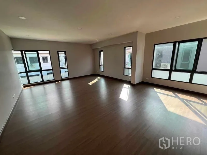 Detached House for rent in Khlong Sam Wa, Bangkok - Spacious empty living room with wood flooring and multiple black-framed windows bringing in natural light.