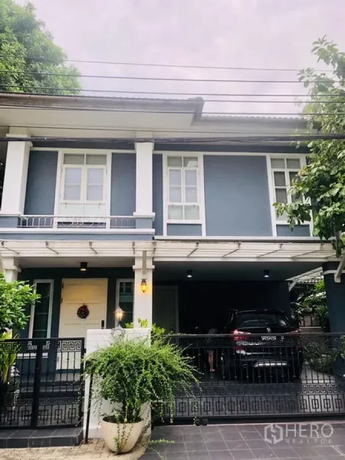 Detached House for rent in Bang Kapi, Bangkok - Two-storey detached house with blue-gray facade, white trim, gated driveway and covered carport surrounded by greenery.