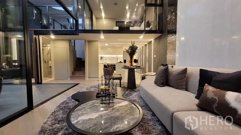 Detached House for sale in Watthana, Bangkok - Spacious lounge with plush rug, glass partitions, and an illuminated open kitchen.