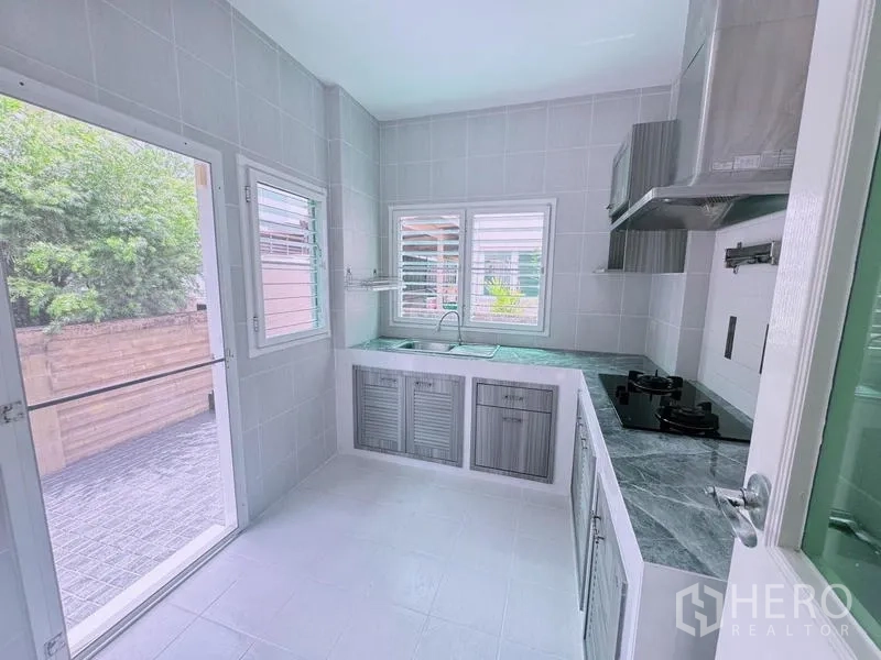 Detached House for rent in Prawet, Bangkok - Bright tiled kitchen with L-shaped counters, gas hob, stainless hood, and windows opening to the patio.