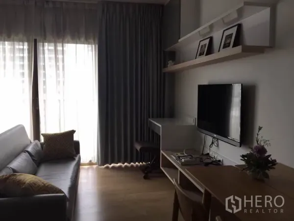 公寓 出租或出售在Bangkok - 客厅配沙发、壁挂电视与书桌，落地窗与窗帘营造明亮舒适的采光。