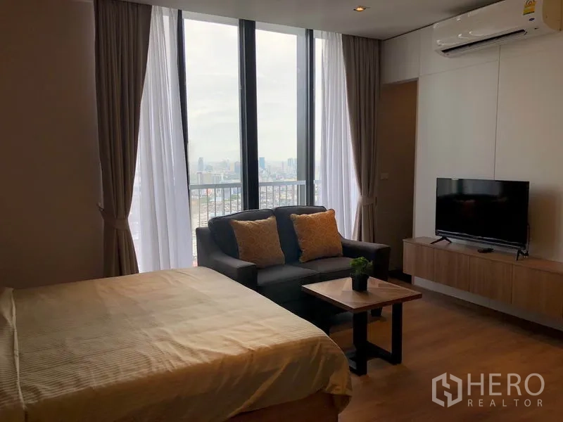 公寓 出租在Khlong Toei, Bangkok - 房间通过高窗欣赏城市景观，床旁有沙发与壁挂电视。