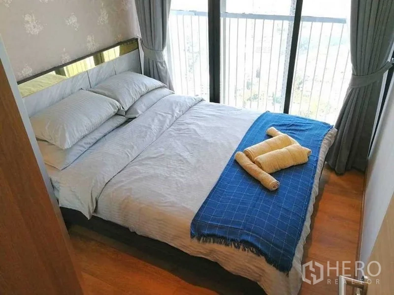 คอนโด ให้เช่า - ห้องนอนขนาดกะทัดรัด เตียงคู่ ผ้าคลุมสีน้ำเงิน และหน้าต่างสูงมองเห็นวิวเมือง