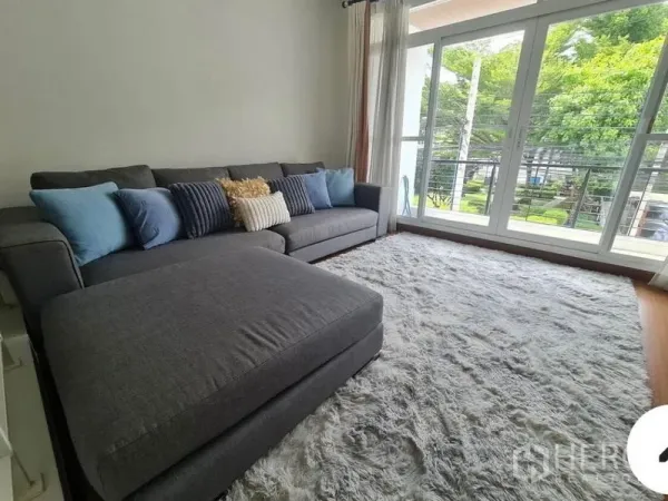 联排别墅 出租在Prawet, Bangkok - 起居室配大灰色沙发、厚地毯，落地窗通向阳台与绿景。