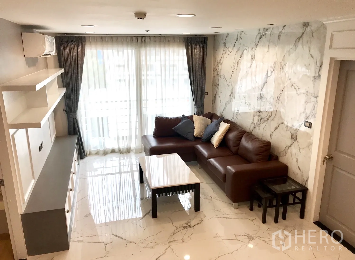 公寓 出租或出售在Khlong Toei, Bangkok - 采光充足的客厅，L型棕色皮沙发、仿大理石地面与落地窗帘。