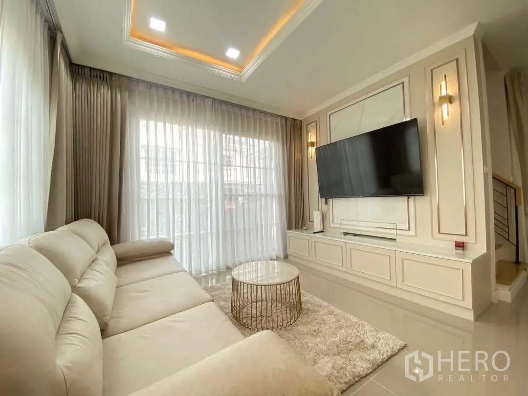 Detached House for rent in Mueang Samut Prakan, Samut Prakan - Living room with L-shaped sofa, TV wall, and floor-to-ceiling curtains.
