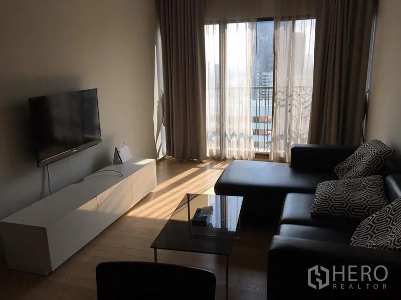 คอนโด ให้เช่าในBangkok - มุมห้องนั่งเล่นพร้อมโซฟารูปตัวแอล ตู้วางทีวี และประตูกระจกเลื่อนออกสู่ระเบียงวิวเมือง