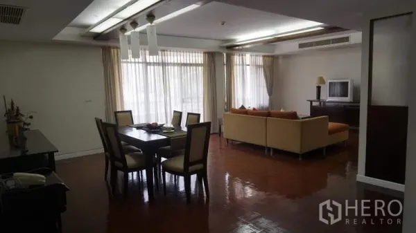 Apartment for rent in Khlong Toei, Bangkok - Bright open-plan living and dining room with large windows, sectional sofa, and a six-seat table on polished wood floors in a Khlong Toei apartment.