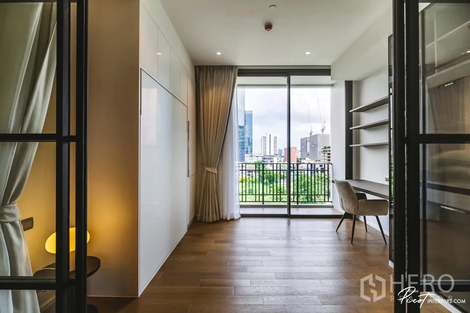 Condominium for rent in Pathum Wan, Bangkok - Bright room with floor-to-ceiling window, balcony facing city and greenery, built-in desk and shelves.