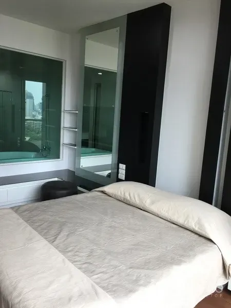 Condominium for rent in Pathum Wan, Bangkok - Bright bedroom with beige bedding, glass window to bathroom featuring a bathtub and a distant city view.