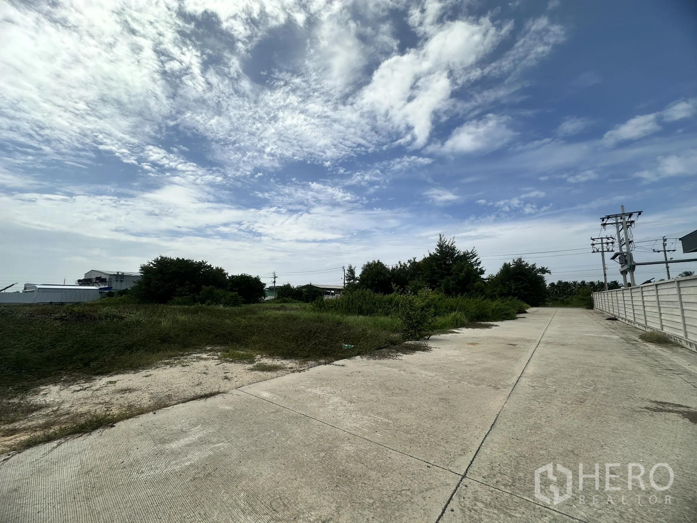 Land for sale in Sai Noi, Nonthaburi - Open land along a wide concrete lane with utility poles and trees under a cloudy sky.