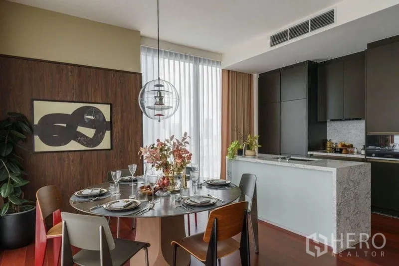 公寓 出租在Watthana, Bangkok - 餐区摆放圆桌，旁为配有大理石中岛与哑光柜体的时尚厨房。