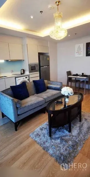 Condominium for rent in Watthana, Bangkok - Open-plan living room with gray sofa, chandelier, rug and compact kitchen beside a dining table.