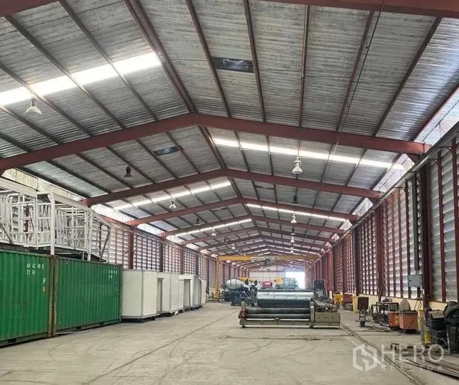 Factory for rent in Bang Kruai, Nonthaburi - Spacious factory interior with high steel truss roof, skylights, louvered walls and concrete flooring with equipment along the sides.