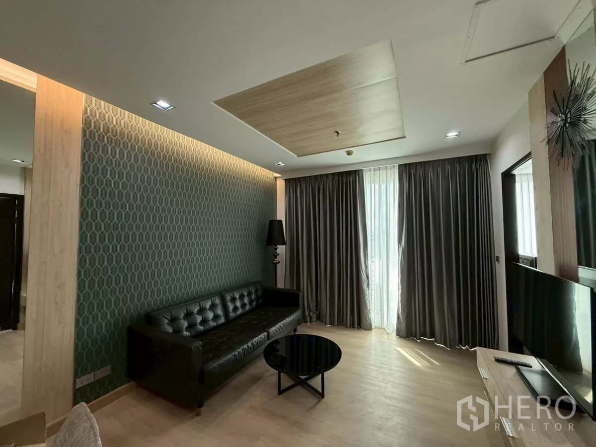 คอนโด ให้เช่าในWatthana, Bangkok - ห้องนั่งเล่นมีผนังลายเด่น โซฟาหนังสีดำ และผ้าม่านสูงจรดเพดาน