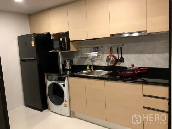 Condominium for rent or sale in Yan Nawa, Bangkok - Fitted kitchen with light wood cabinets, black stone countertop, fridge, microwave, and washing machine.
