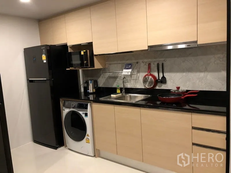 Condominium for rent or sale in Yan Nawa, Bangkok - Fitted kitchen with light wood cabinets, black stone countertop, fridge, microwave, and washing machine.