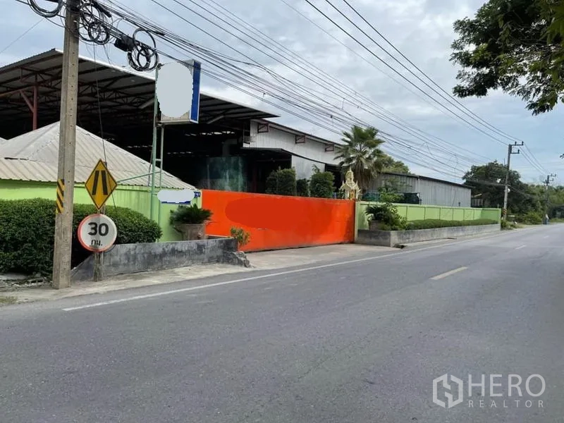 货栈 出售在Lam Luk Ka, Pathum Thani - 仓库正面斜视图，橙色推拉门、路边指示牌与宽阔钢棚顶。