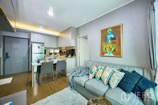 Condominium for rent in Phra Khanong, Bangkok - Living area with sofa, rug and artwork looking toward the compact kitchen and dining zone in a modern condo.