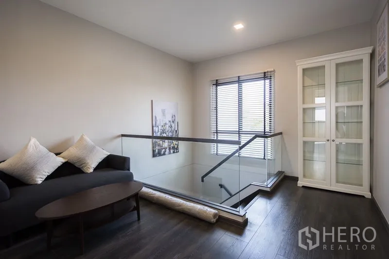 联排别墅 出租在Samut Prakan - 二层楼梯平台休闲角，玻璃护栏、沙发与展示柜。