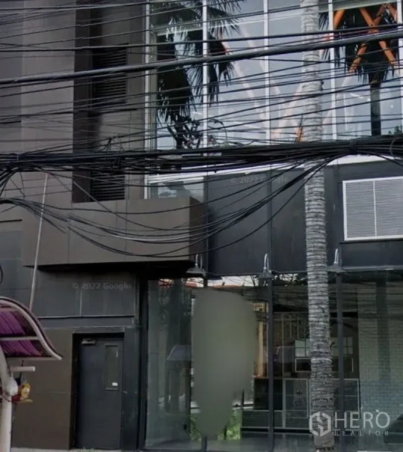 零售空间 出租在Watthana, Bangkok - 街道视角的现代深色玻璃店铺外立面，落地窗、棕榈树与上方电缆。