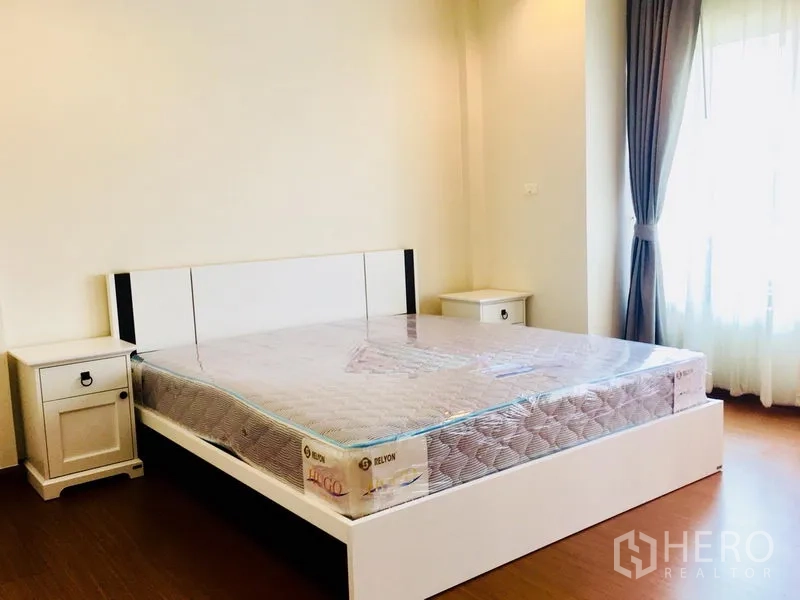 Townhouse for rent in Prawet, Bangkok - Second bedroom with a large bed, bedside tables, curtained window, and wood floor.