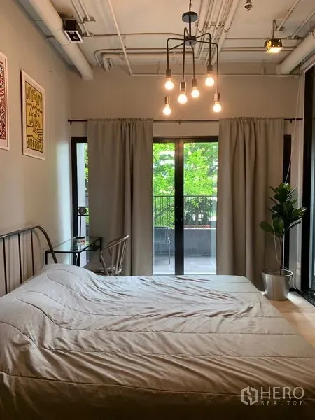 Condominium for sale in Watthana, Bangkok - Cozy bedroom with exposed ceiling lights, sliding doors to the balcony, curtains, and a potted plant.