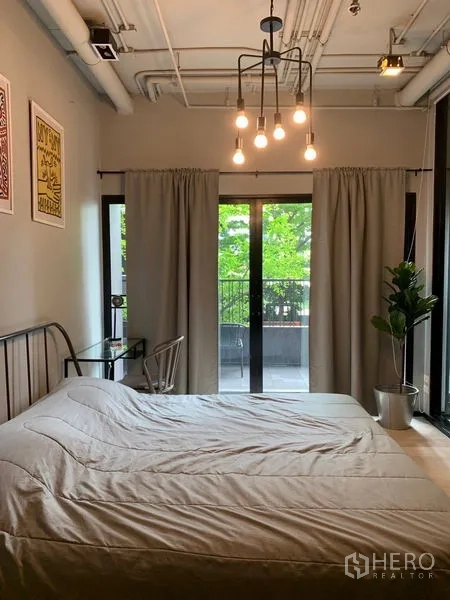 Condominium for sale in Watthana, Bangkok - Cozy bedroom with exposed ceiling lights, sliding doors to the balcony, curtains, and a potted plant.