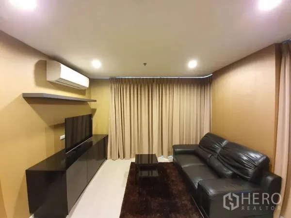 公寓 出租在Sathon, Bangkok - 客厅内黑色皮沙发、电视矮柜、置物层板与棕色地毯，射灯照明。