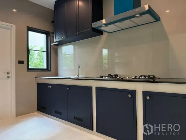 Detached House for rent or sale in Bang Phli, Samut Prakan - Contemporary kitchen with navy cabinetry, gas hob, chimney hood and a window bringing in garden views.