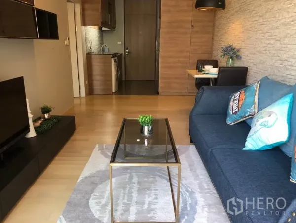 คอนโด ให้เช่าในBangkok - ห้องนั่งเล่นสไตล์โมเดิร์น โซฟาสีน้ำเงิน โต๊ะกระจก ชั้นวางทีวี และครัวเปิดพร้อมเครื่องซักผ้าในคอนโดกรุงเทพฯ