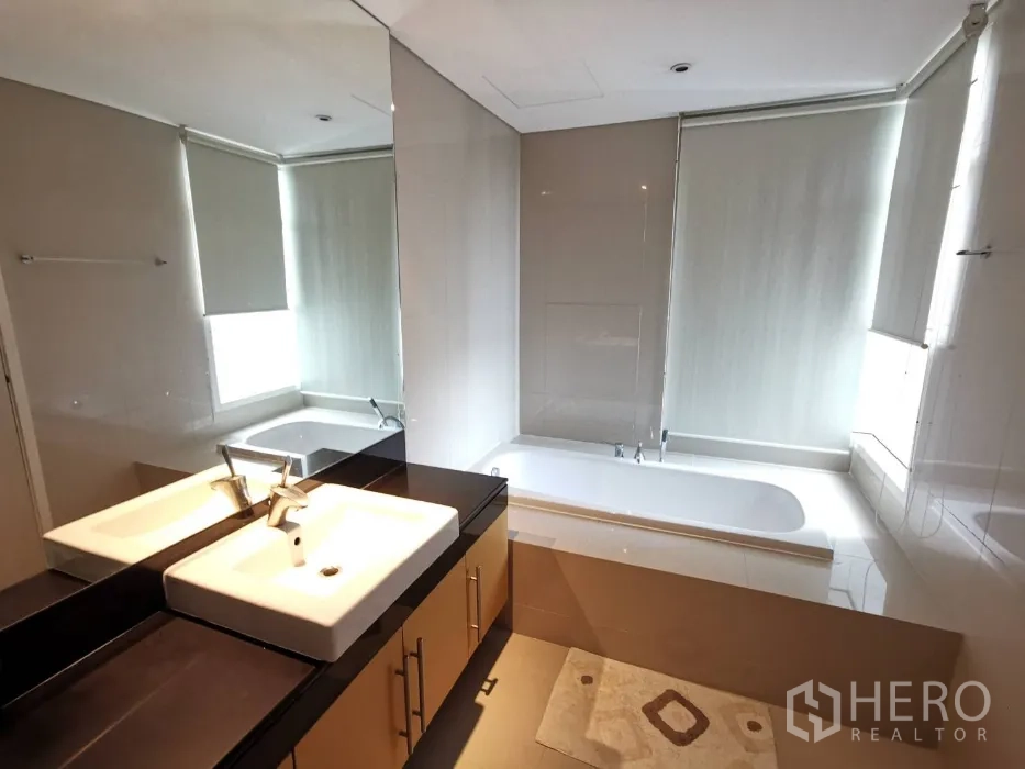 Condominium for rent in Watthana, Bangkok - Contemporary bathroom with long vanity, large mirror, and a built-in bathtub beside a window with blinds.