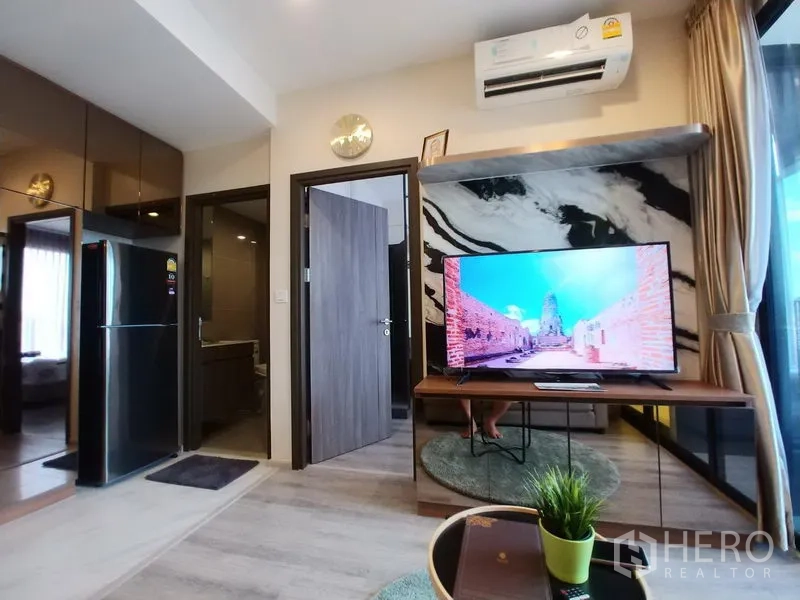 Condominium for rent in Huai Khwang, Bangkok - Living room with marble accent wall and TV, air conditioner above, and doors to the bedroom and bathroom.