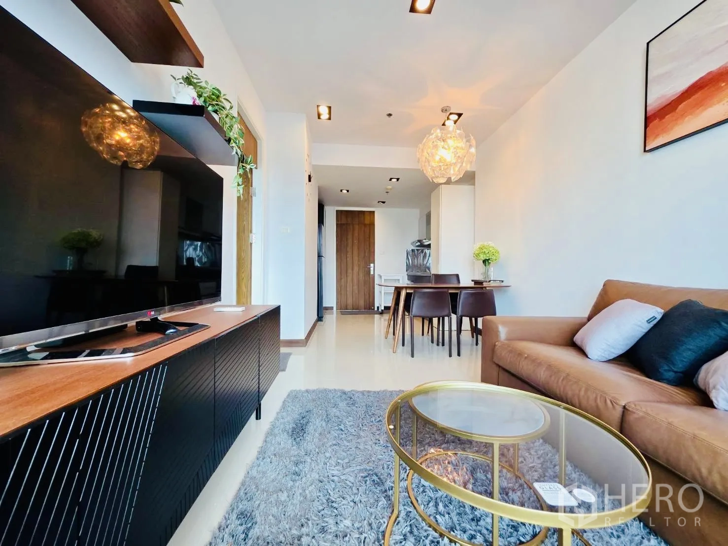 Condominium for rent in Huai Khwang, Bangkok - Open-plan lounge with TV console and shelves facing a brown leather sofa and dining set.