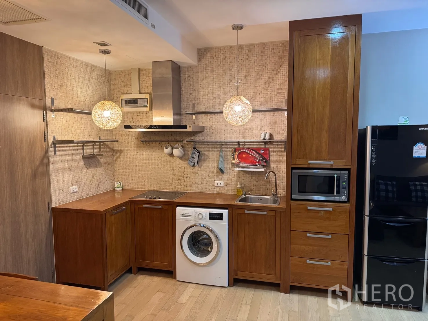 คอนโด for rent or sale in Watthana, Bangkok - ครัวแบบเปิดตู้ไม้ เตาไฟฟ้า เครื่องดูดควัน อ่างล้างจาน ไมโครเวฟ ตู้เย็น และเครื่องซักผ้า ใต้โคมไฟแขวน