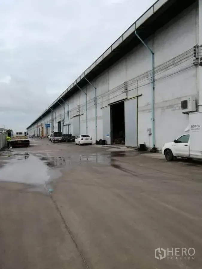 货栈 出租在Samut Prakan - 一排工业仓库外立面，湿滑混凝土道路上停有车辆和装卸区。