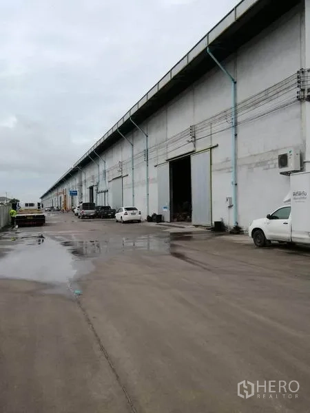 货栈 出租在Samut Prakan - 一排工业仓库外立面，湿滑混凝土道路上停有车辆和装卸区。