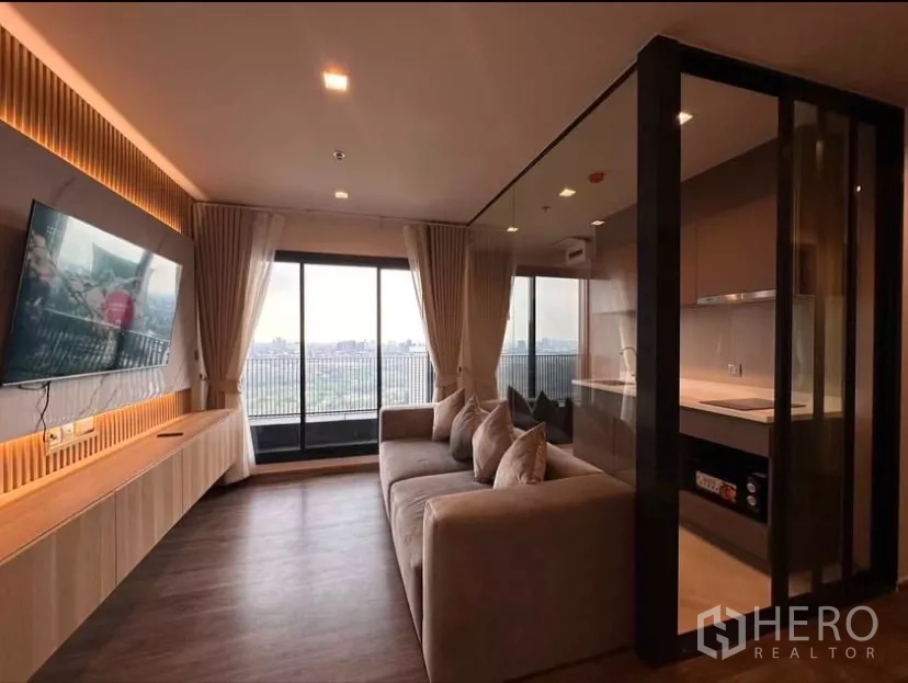 Condominium for rent in Chatuchak, Bangkok - Living area with wall-mounted TV, large sofa, and panoramic city view from the balcony.