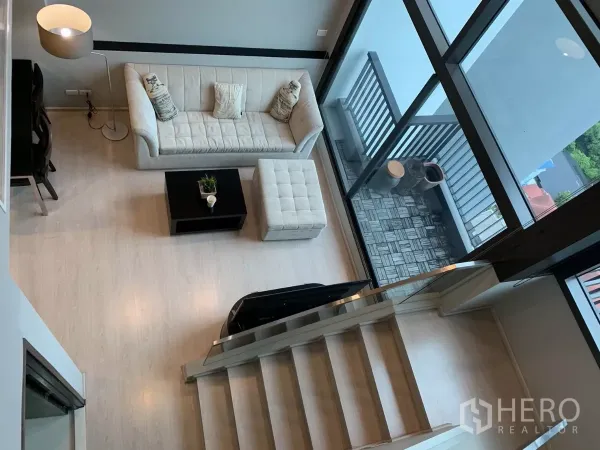 คอนโด ให้เช่าในKhlong Toei, Bangkok - มุมสูงของห้องนั่งเล่นเพดานสูง โซฟาสีครีม บันได และระเบียงหลังหน้าต่างกระจกเต็มผนัง