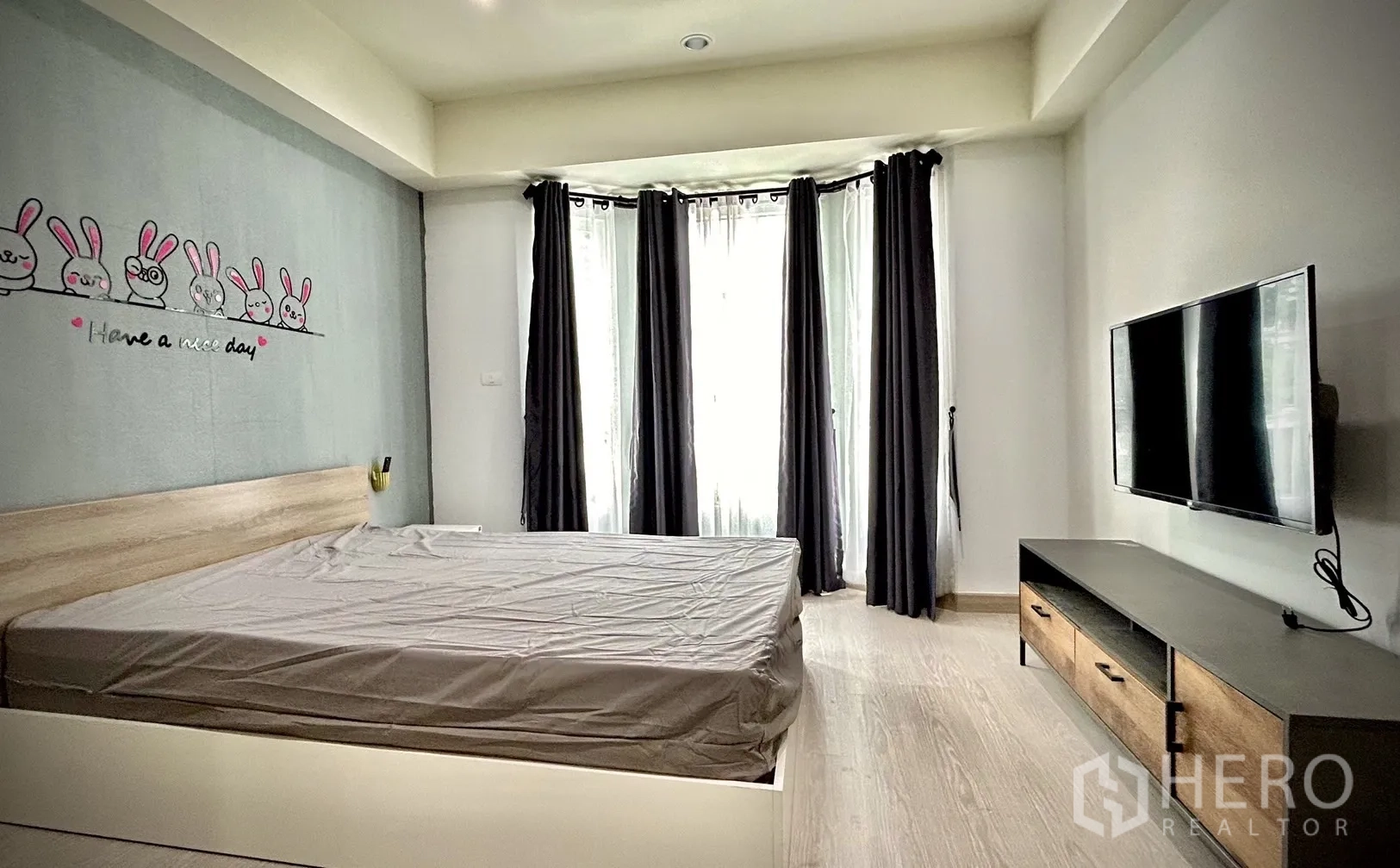 Townhouse for rent in Bang Kapi, Bangkok - Second bedroom with bay windows, wall-mounted TV and wood-and-metal TV cabinet.
