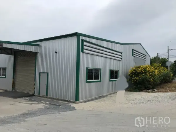 Warehouse for rent in Pak Kret, Nonthaburi - Modern metal-clad warehouse exterior with green trim, roller shutter door and side access beside landscaped shrubs.