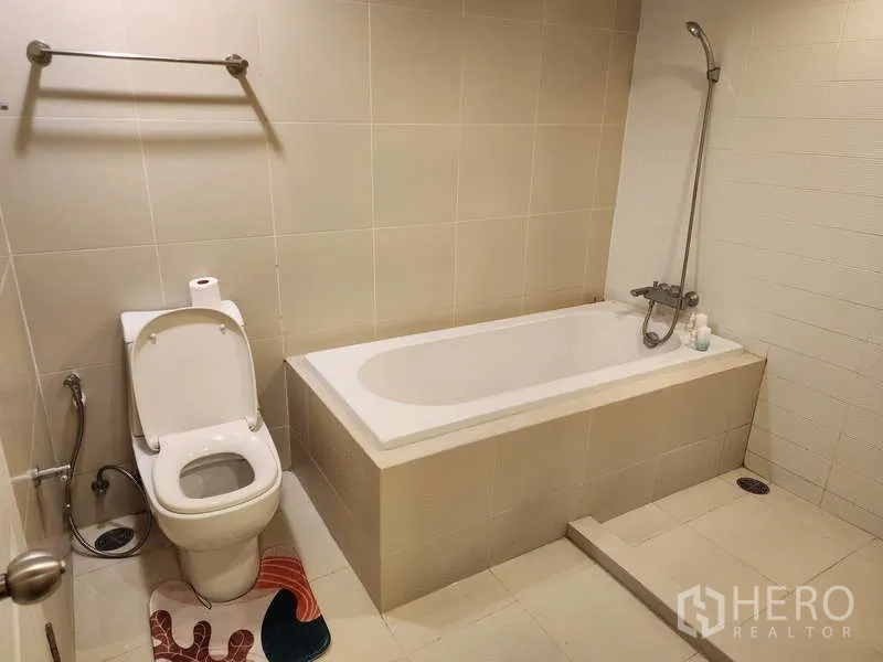 Condominium for rent in Huai Khwang, Bangkok - Bathroom with built-in bathtub, handheld shower and toilet finished in beige tiles.