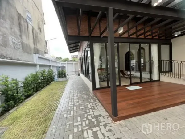 Detached House for rent in Ratchathewi, Bangkok - Covered wooden deck beside a glass-walled living room and a paved side walkway.