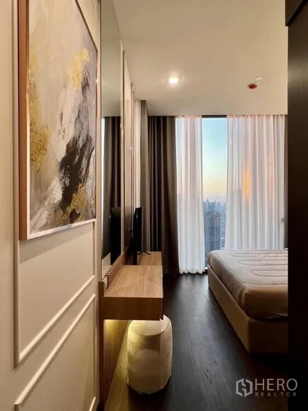 Condominium for rent in Watthana, Bangkok - Bedroom with built-in desk and TV facing skyline views through full-height curtains at dusk.