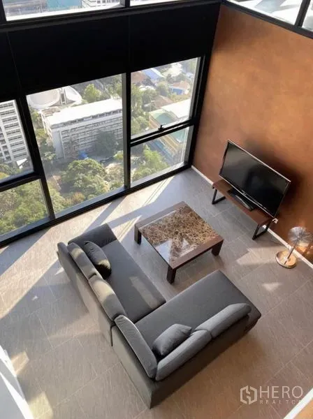 公寓 出租在Bang Rak, Bangkok - 俯视客厅：L形沙发、大理石茶几与电视，临近落地窗眺望天际线。