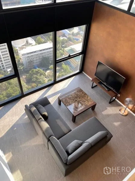 公寓 出租在Bang Rak, Bangkok - 俯视客厅：L形沙发、大理石茶几与电视，临近落地窗眺望天际线。