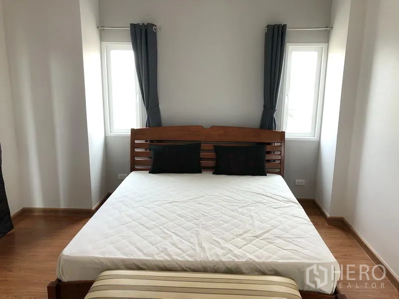 Detached House for rent or sale in Huai Khwang, Bangkok - Corner bedroom with wooden bedframe, twin windows and neutral curtains.