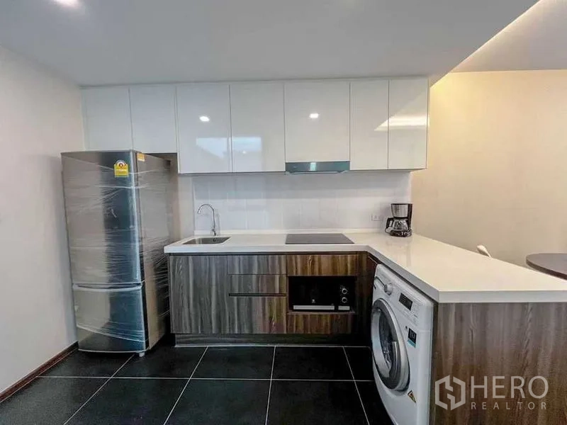 Condominium for sale in Huai Khwang, Bangkok - L-shaped kitchen with glossy cabinets, electric hob, oven, refrigerator and a front-loading washing machine.
